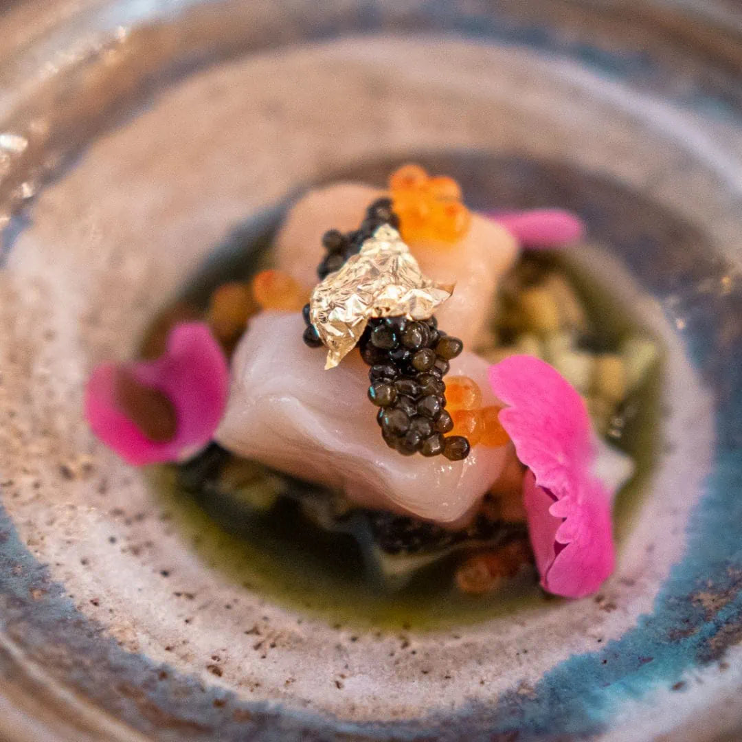 Delicate dish with caviar, pink flowers, seaweed and a gold leaf on a textured plate.